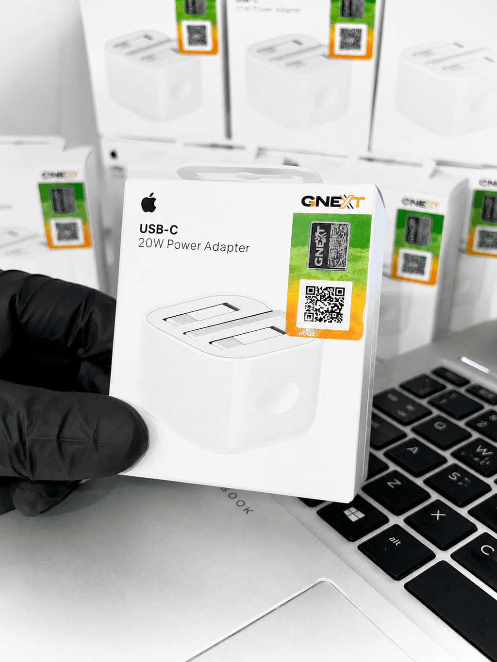 Apple USB-C 20W Power Adapter in retail packaging held over a laptop keyboard.