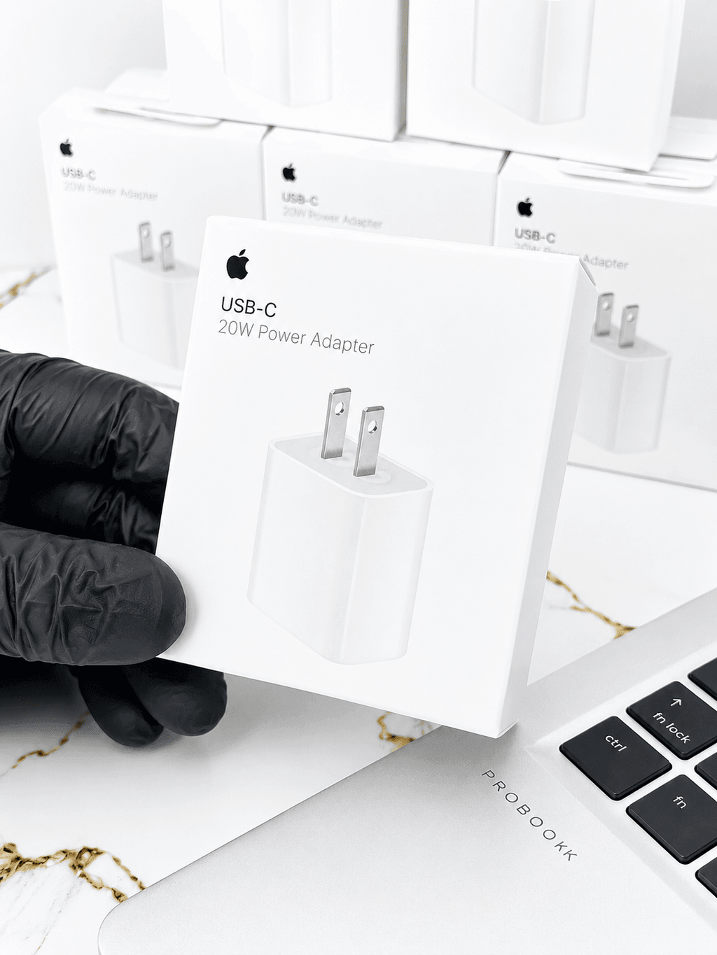Hand holding an Apple USB-C 20W Power Adapter box against a background of multiple adapter boxes.