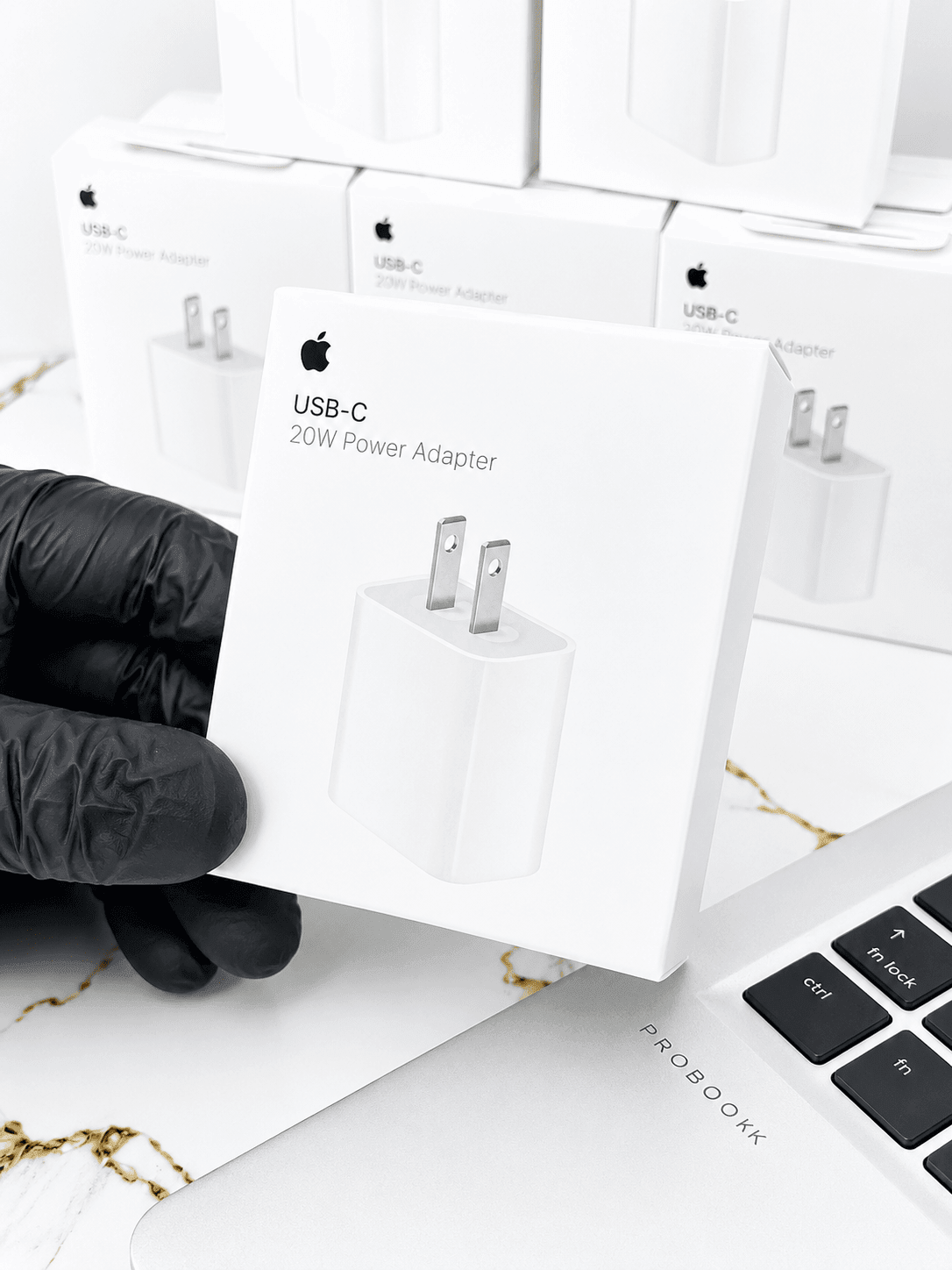 Hand holding an Apple USB-C 20W Power Adapter box against a background of multiple adapter boxes.