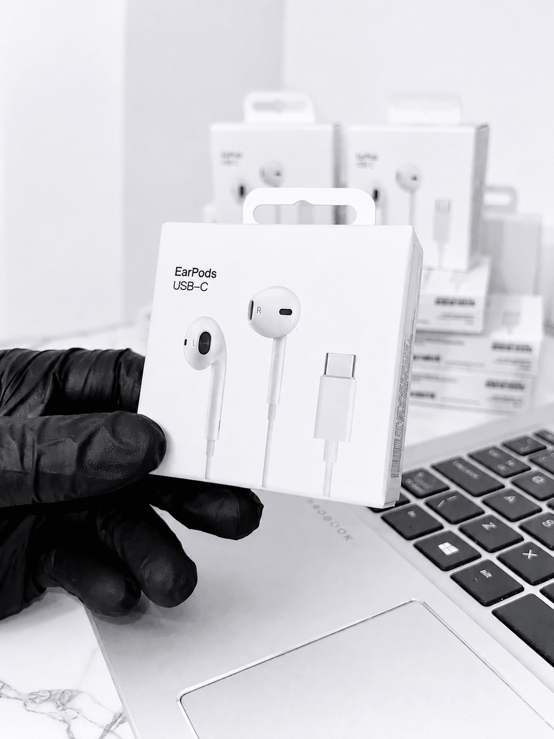 A hand in a black glove holding a box of USB-C EarPods next to a laptop.